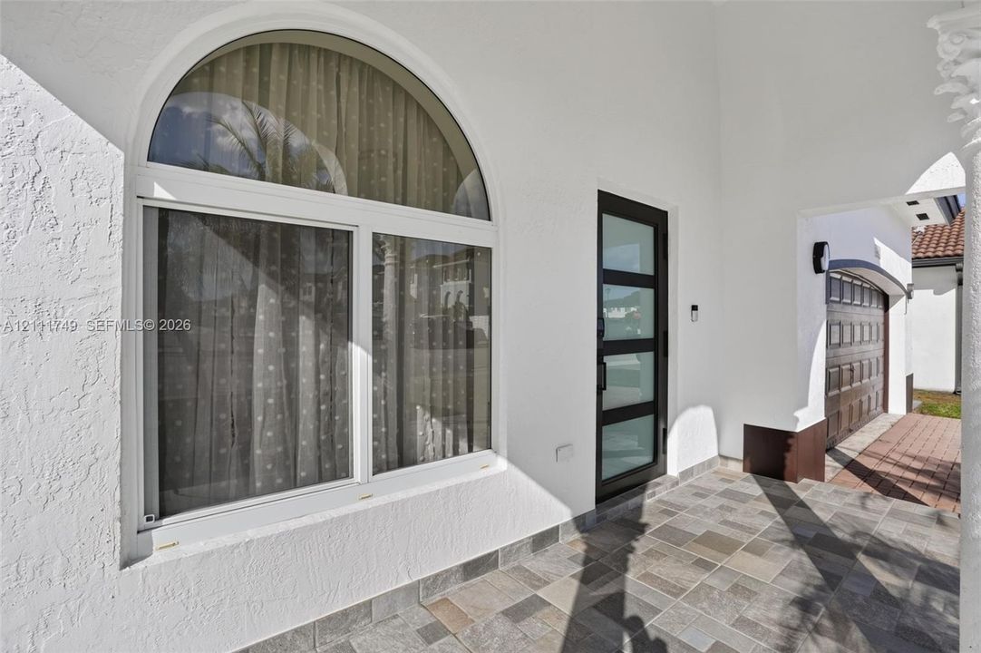 Covered entryway featuring multi-tone tile flooring, arched window with white framing, modern glass-paneled entry door, and architectural column detail