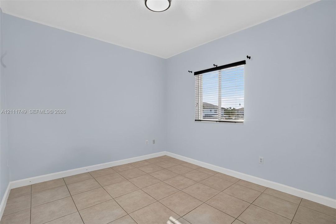 Neutral-toned interior space featuring light-colored wall paint, ceramic tile flooring, and a window with horizontal blinds and an attached curtain rod