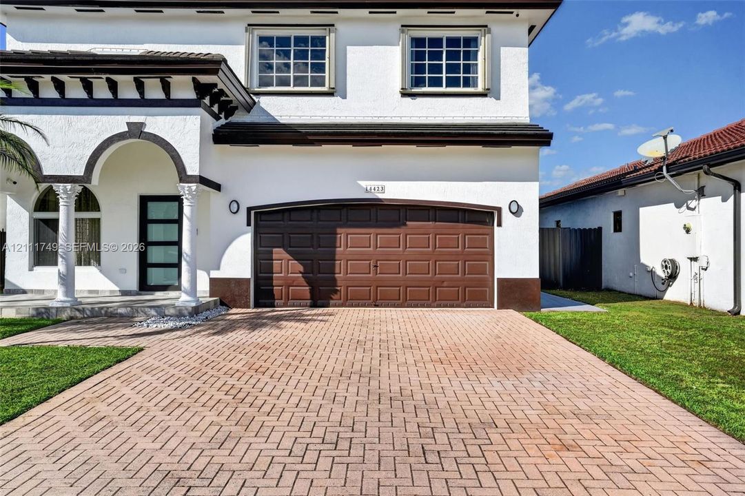 Two-story stucco exterior featuring a tiled roof, arched covered entryway with decorative columns, paver driveway, and an attached multi-panel garage door