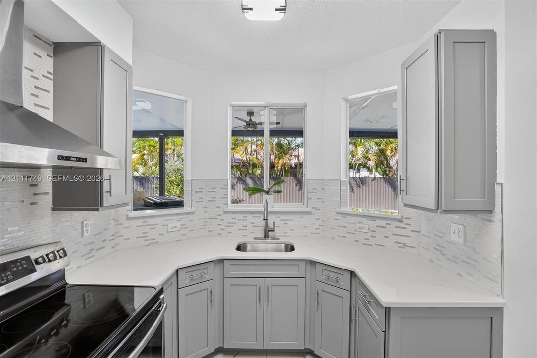 Kitchen featuring grey shaker-style cabinetry, quartz countertops, a stainless steel undermount sink with a gooseneck faucet, a patterned tile backsplash, and a stainless steel range hood