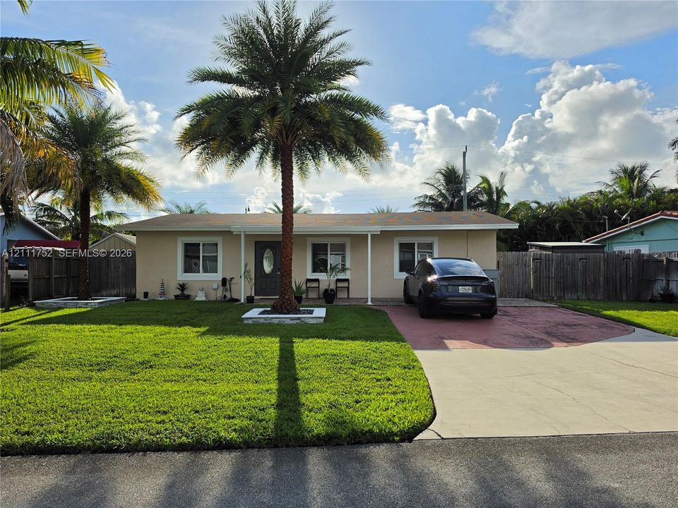 Single-story residence featuring a stucco exterior, concrete driveway, front yard landscaping, and mature palm trees enclosed by wooden perimeter fencing