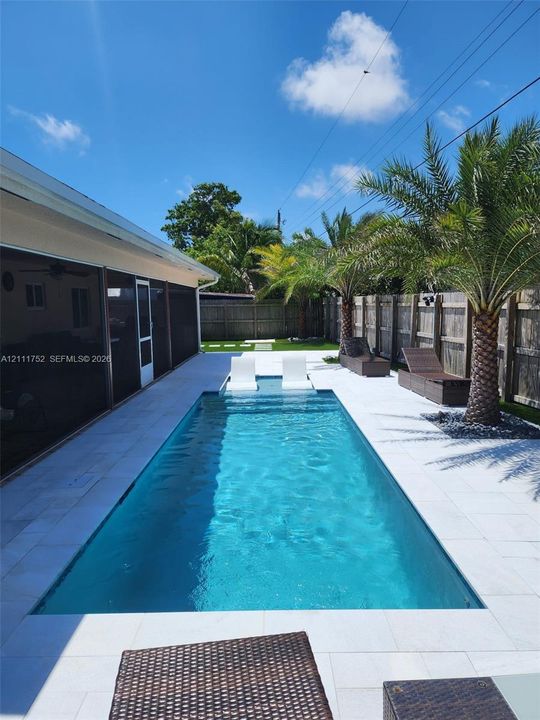 Rectangular swimming pool featuring a Baja shelf, light-toned stone pool deck, and perimeter wood fencing accented by mature palm trees