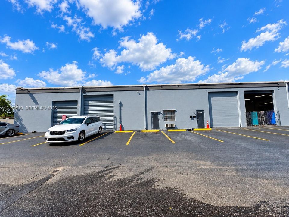Single-story industrial exterior featuring a smooth gray concrete facade, three metal roll-up doors, two dedicated entry points, and an asphalt-paved parking lot with marked stalls