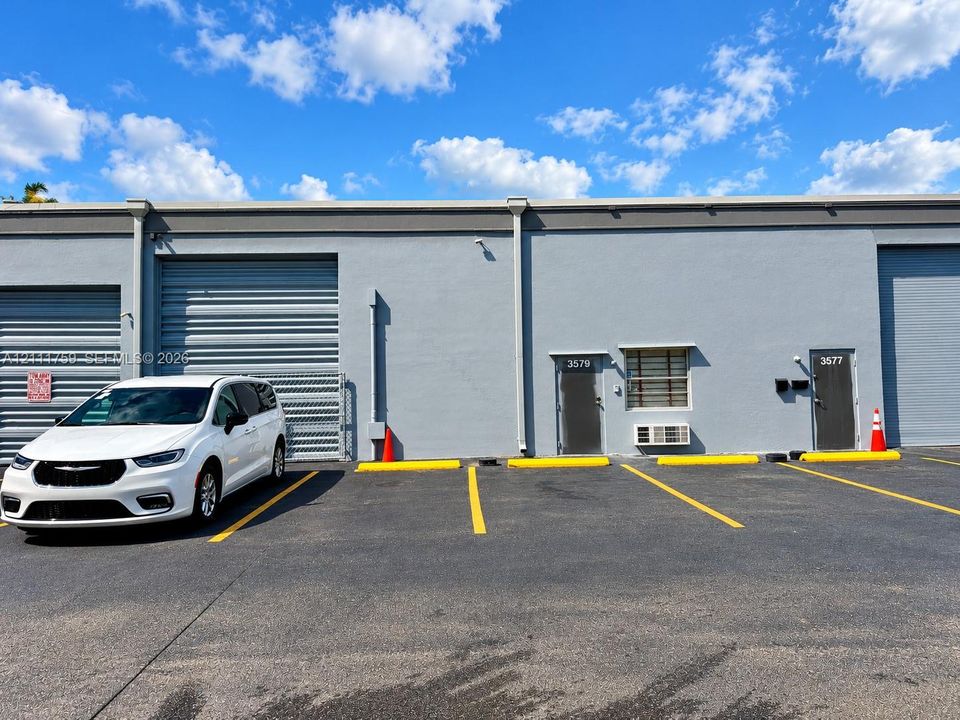 Exterior commercial facade featuring gray concrete construction, multiple metal roll-up bay doors, individual pedestrian entry doors, and dedicated asphalt parking spaces with yellow markings