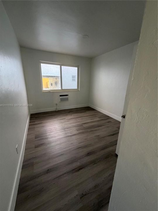 Neutral-toned interior room featuring wood-finish flooring, white walls, and a window with an integrated wall-mounted air conditioning unit