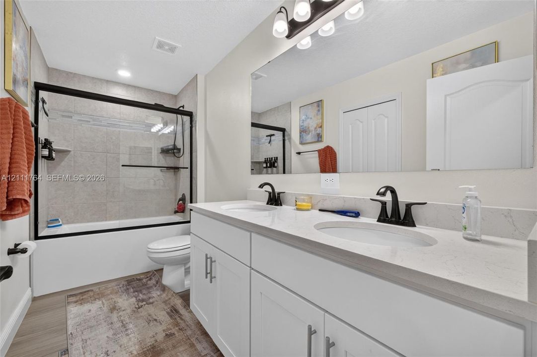 Dual-sink vanity with white cabinetry, light-toned stone-finish countertop, and matte black hardware, paired with a glass-enclosed shower featuring neutral tile surround and matte black fixtures