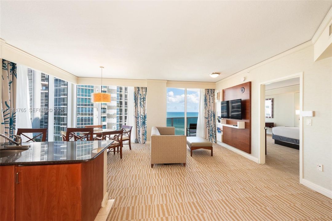 Open-concept living space featuring floor-to-ceiling windows, built-in wooden media cabinetry, a kitchen island with stone countertop, and a private balcony with water scenery