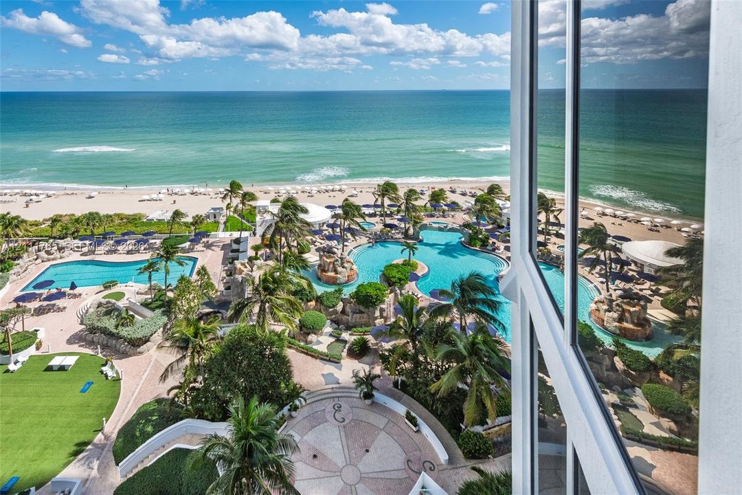 High-rise perspective featuring floor-to-ceiling windows, outdoor swimming pools, manicured green space, stone-paved plaza, and oceanfront exposure