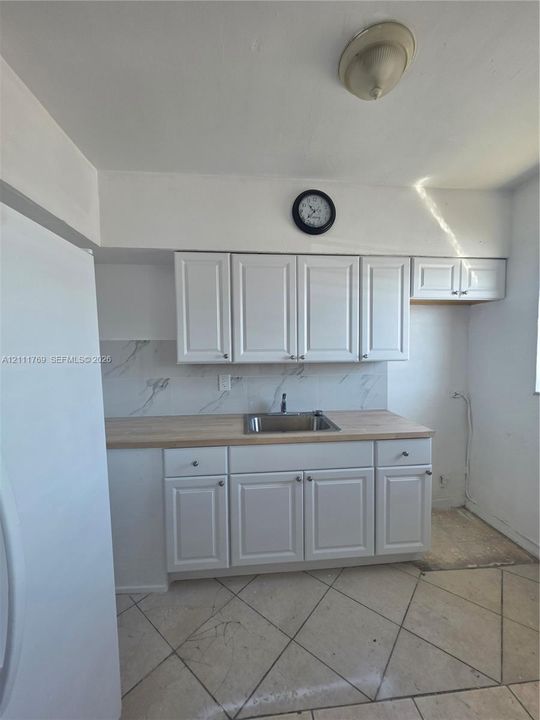 Kitchen workspace featuring white shaker-style cabinetry, wood-finish countertops, stone-look backsplash, stainless steel drop-in sink, and ceramic tile flooring