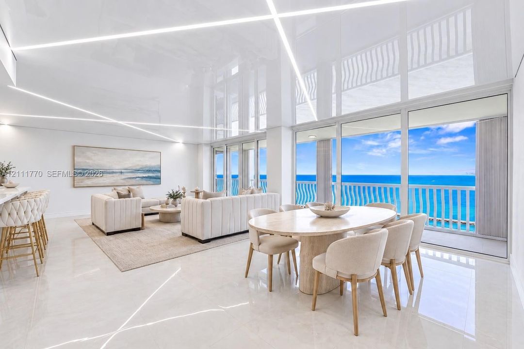 High-gloss reflective ceiling with integrated linear LED lighting, polished stone flooring, and expansive floor-to-ceiling glass sliding doors opening to an oceanfront balcony with structural railing