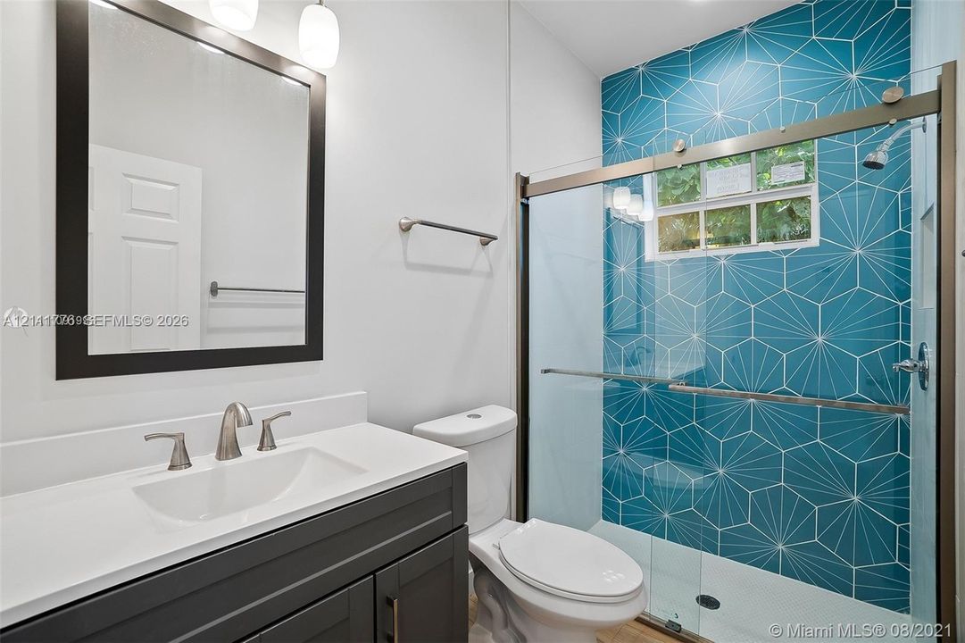 Bathroom featuring a charcoal vanity with a white countertop, geometric blue accent tile in the shower, glass shower enclosure, and a rectangular mirror with a dark frame