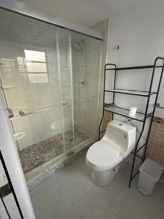 Walk-in shower featuring a sliding glass enclosure, pebble-stone flooring, and a wall-mounted showerhead, complemented by a dual-flush toilet and neutral tile wall accents
