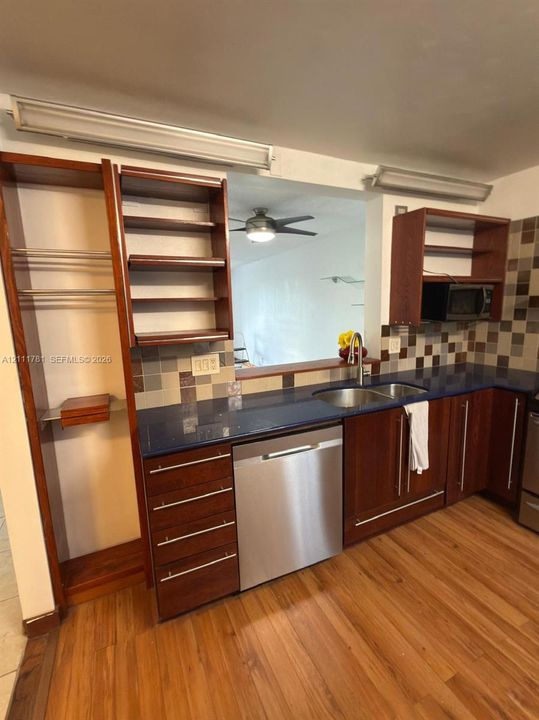 Kitchen featuring wood cabinetry with built-in open shelving, dark countertops, a double-basin stainless steel sink, stainless steel dishwasher, and multi-toned tile backsplash