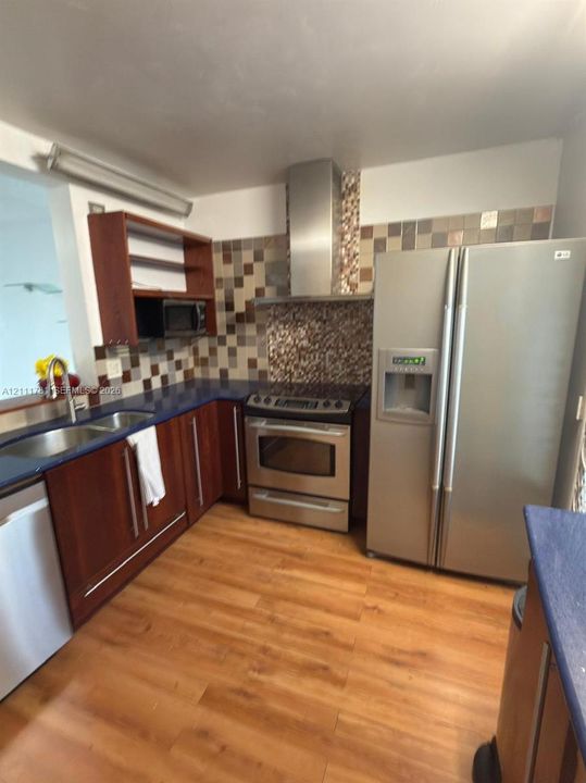 Kitchen featuring dark-toned cabinetry, stainless steel appliances, a chimney-style range hood, decorative tile backsplash, and wood-finish flooring
