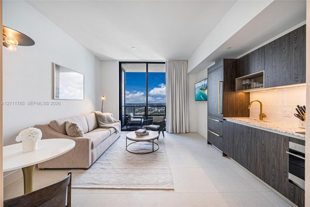 Kitchen featuring dark wood-finish cabinetry, under-cabinet lighting, stone-surface countertops, and gold-toned faucet hardware, floor-to-ceiling glass doors opening to a balcony with an expansive urban backdrop