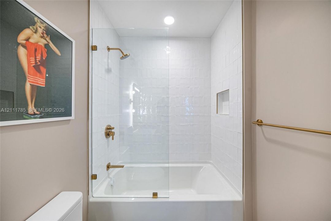 Bathtub and shower combination featuring a glass panel door, white tile surround, integrated shelving niche, and brass-finish fixtures
