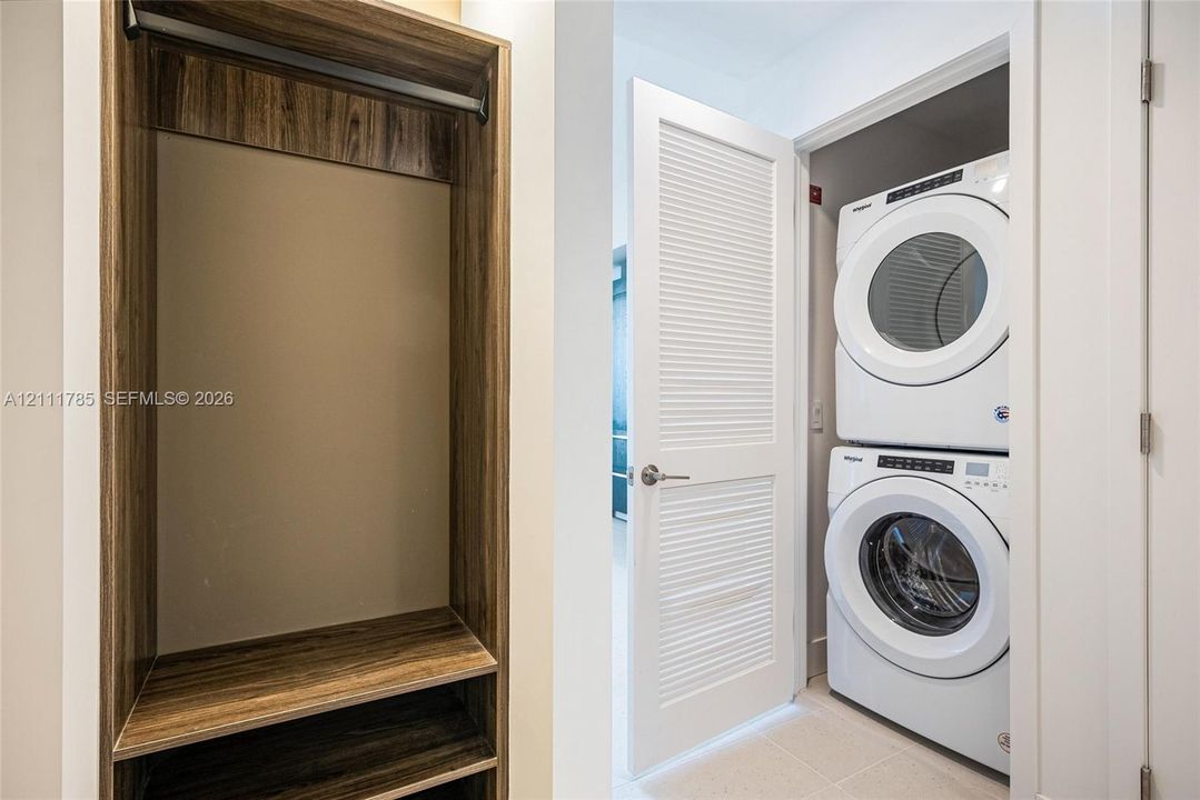 Built-in storage cabinet with wood-finish shelving and hanging rod, paired with a louvered door providing access to a stacked washer and dryer unit