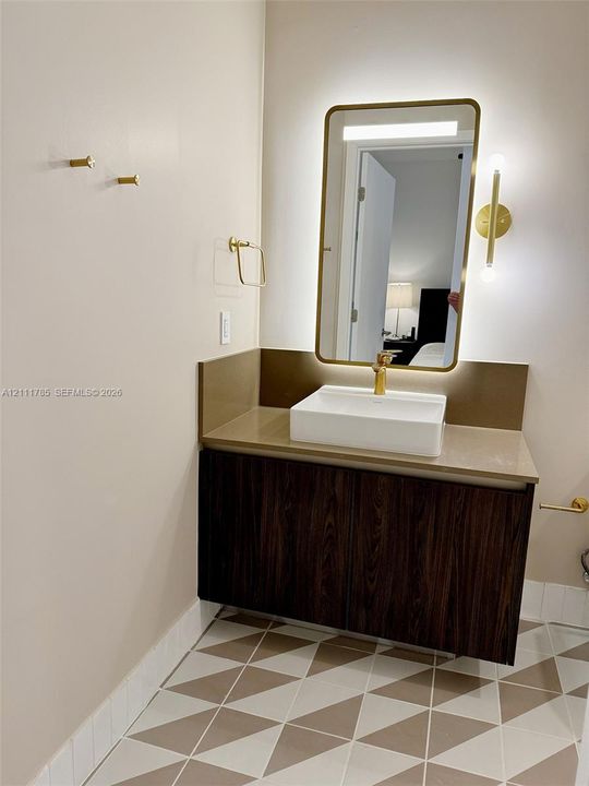 Floating vanity with wood-finish cabinetry, rectangular vessel sink, brass-tone hardware, backlit wall mirror, and geometric patterned floor tiling