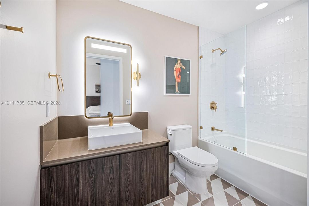 Floating vanity with stone-surface countertop, rectangular vessel sink, brass-finish fixtures, LED-backlit mirror, and glass-enclosed shower with white wall tiling