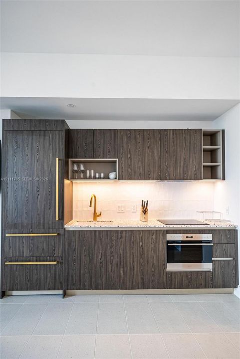Dark wood-finish cabinetry, brass-toned hardware, stone-composite countertop, integrated refrigeration, and stainless steel wall oven