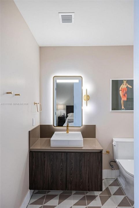 Vanity featuring dark wood-finish cabinetry, a solid surface countertop, rectangular vessel sink, and brass-tone fixtures, complemented by a backlit rectangular mirror and geometric tile flooring