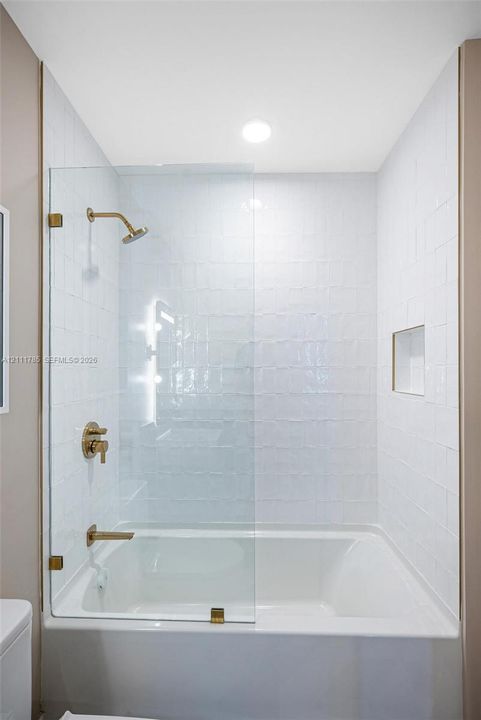 Bathtub and shower combination featuring white textured wall tile, glass shower panel with gold-tone hardware, recessed shower niche, and gold-tone wall-mounted faucet and shower head