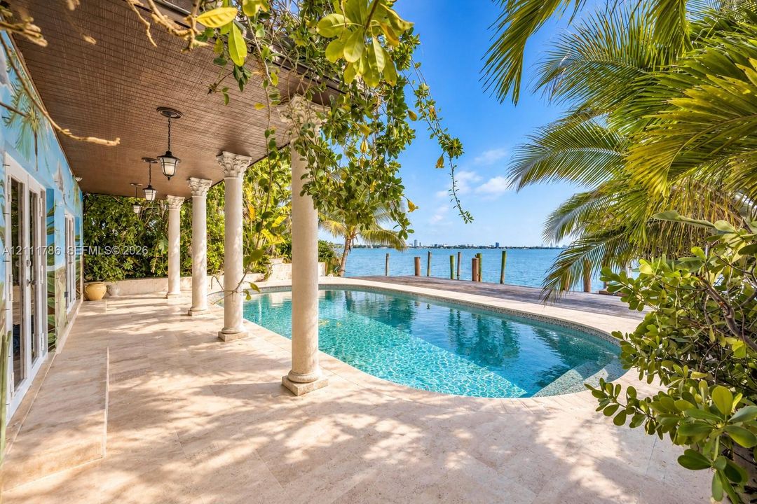 Covered stone patio featuring classical columns, integrated lantern-style pendant lighting, and a kidney-shaped pool with stone decking