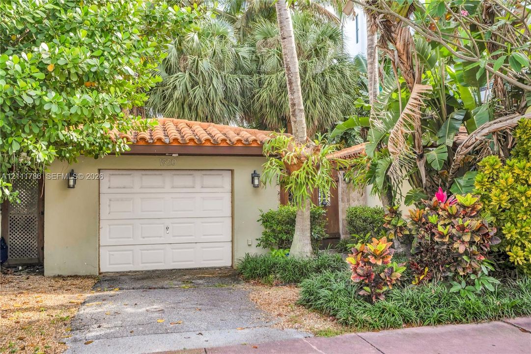 Single-car garage featuring a terracotta tile roof, stucco exterior, and wall-mounted coach lighting, surrounded by mature palm trees and tropical landscaping