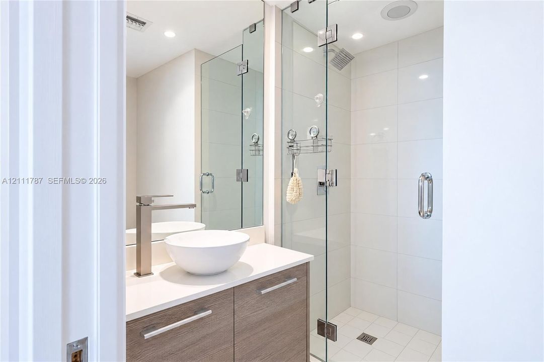 Bathroom featuring a wood-finish vanity with a white countertop, vessel sink, polished metal faucet, and a frameless glass-enclosed shower with white wall tile
