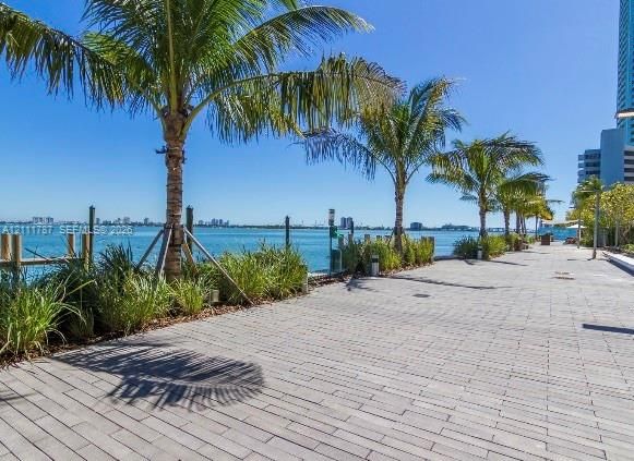 Waterfront walkway featuring concrete paver flooring, mature palm trees, native landscaping, and wooden piling markers