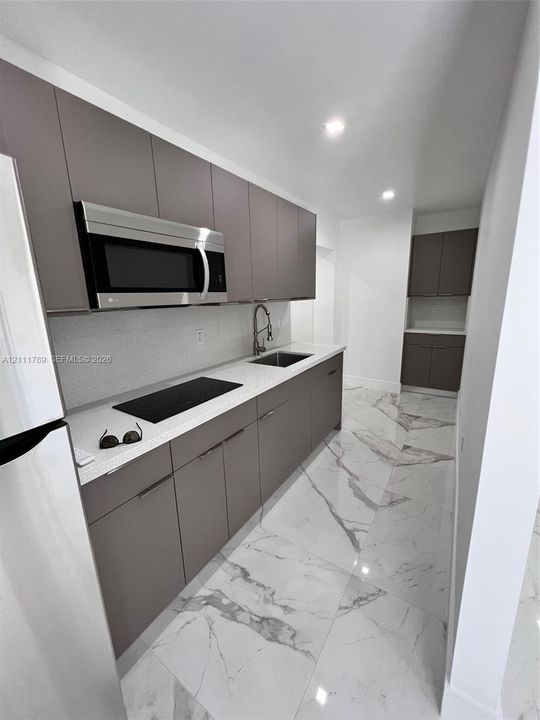 Galley kitchen featuring flat-panel cabinetry, light-toned countertops with an integrated sink, stainless steel microwave, and large-format tile flooring