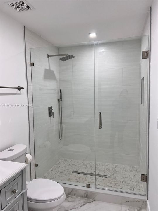 Walk-in shower featuring a frameless glass enclosure, rain-style shower head, handheld sprayer, neutral tiled surround, and stone-mosaic floor tiling