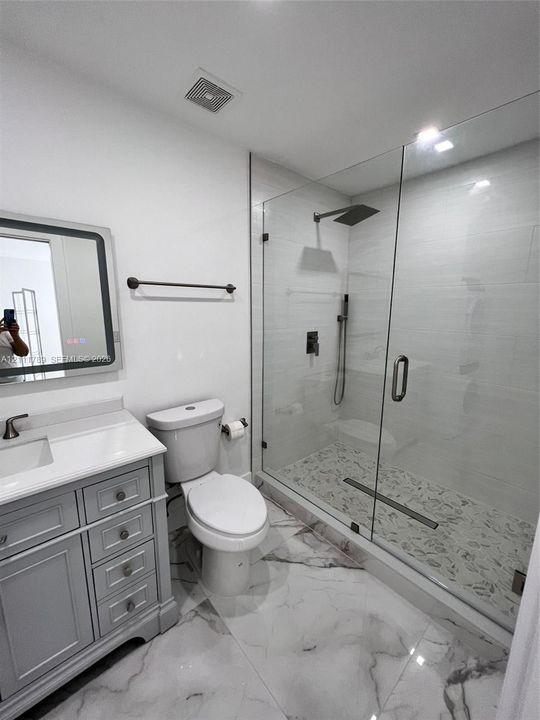 Grey shaker-style vanity with white countertop, frameless glass walk-in shower featuring a rainfall showerhead, pebble-finish shower floor, and large-format polished tile flooring