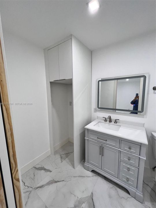 Vanity featuring a stone-composite countertop with an integrated rectangular sink and metallic faucet, soft-grey shaker-style cabinetry with tiered storage, wall-mounted backlit mirror, and large-format porcelain tile flooring with grey veining