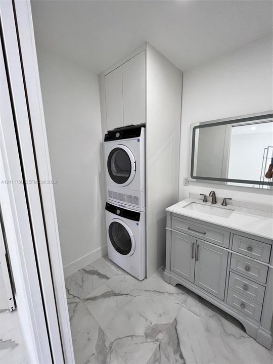 Floor-to-ceiling built-in cabinetry, stacked laundry appliances, vanity with a white countertop and integrated sink, stone-finish tile flooring, and a rectangular mirror with integrated LED lighting