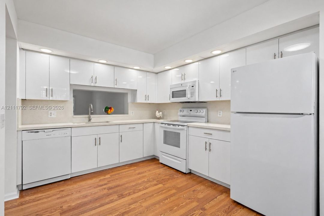 Brand new kitchen, featuring white high-gloss cabinetry, wood-finish flooring, integrated recessed lighting, and a passthrough window. New appliances