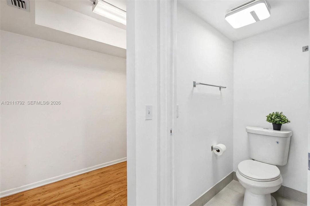 Restroom featuring a dual-flush toilet, tiled flooring, wall-mounted towel bar, and ceiling-mounted LED lighting fixture, with wood-finish flooring in the surrounding area