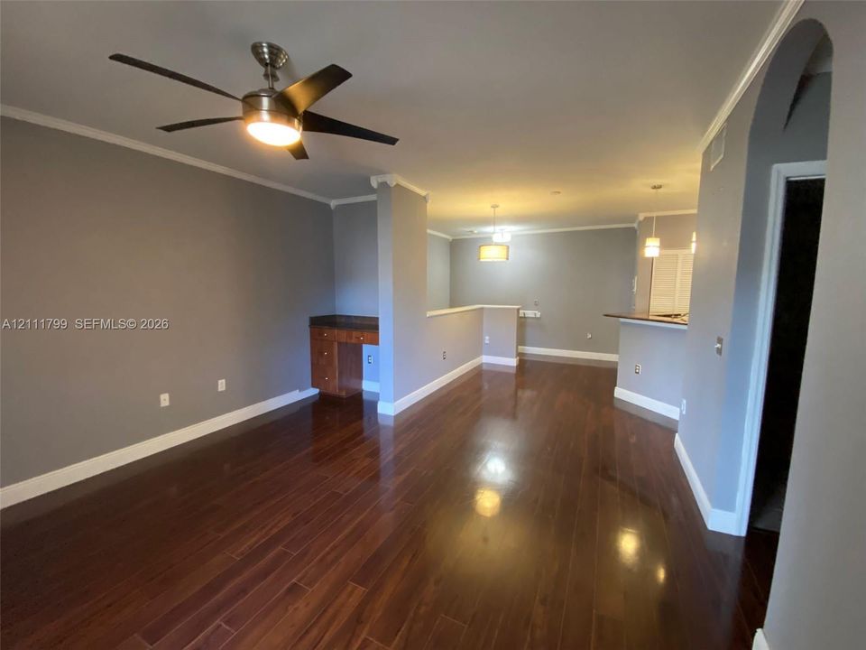 Open-concept interior featuring wood-finish flooring, a built-in wooden desk with a stone countertop, an integrated breakfast bar, crown molding, and a multi-blade ceiling fan with central lighting