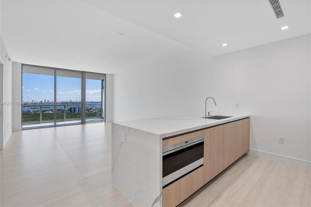 Kitchen island featuring a stone-look waterfall countertop, integrated microwave, light wood-finish cabinetry, and a stainless steel undermount sink with a gooseneck faucet, set against floor-to-ceiling glass panel windows