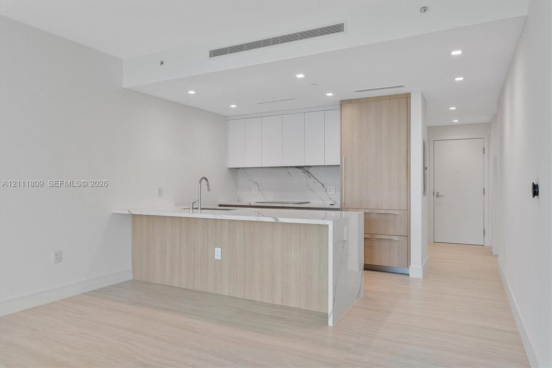 Kitchen featuring flat-panel cabinetry, light-toned wood-finish millwork, stone-patterned countertops, a waterfall island, and integrated panel-ready appliances