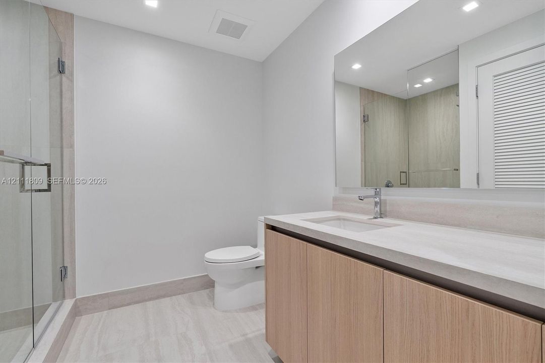 Bathroom featuring a vanity with a light wood-finish cabinet and stone-look countertop, integrated under-mount sink, large wall-mounted mirror, glass-enclosed shower, and tile flooring