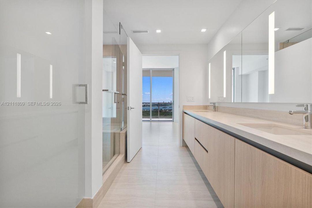 Bathroom featuring a dual-sink vanity with light wood-finish cabinetry, solid surface countertop, large wall-mounted mirrors with integrated vertical lighting, and a glass-enclosed shower
