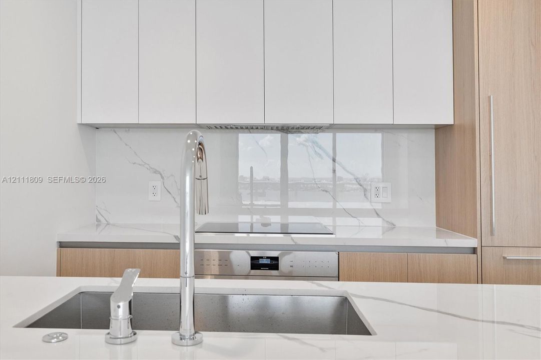 Sleek kitchen featuring flat-panel cabinetry, light-toned wood-finish lower cabinets, stone-surfaced countertops, matching full-height backsplash, and a stainless steel undermount sink with a high-arc pull-down faucet