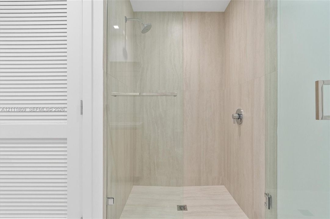 Walk-in shower featuring large-format neutral tile surround, frameless glass enclosure, polished chrome rainfall showerhead, and a built-in shelf