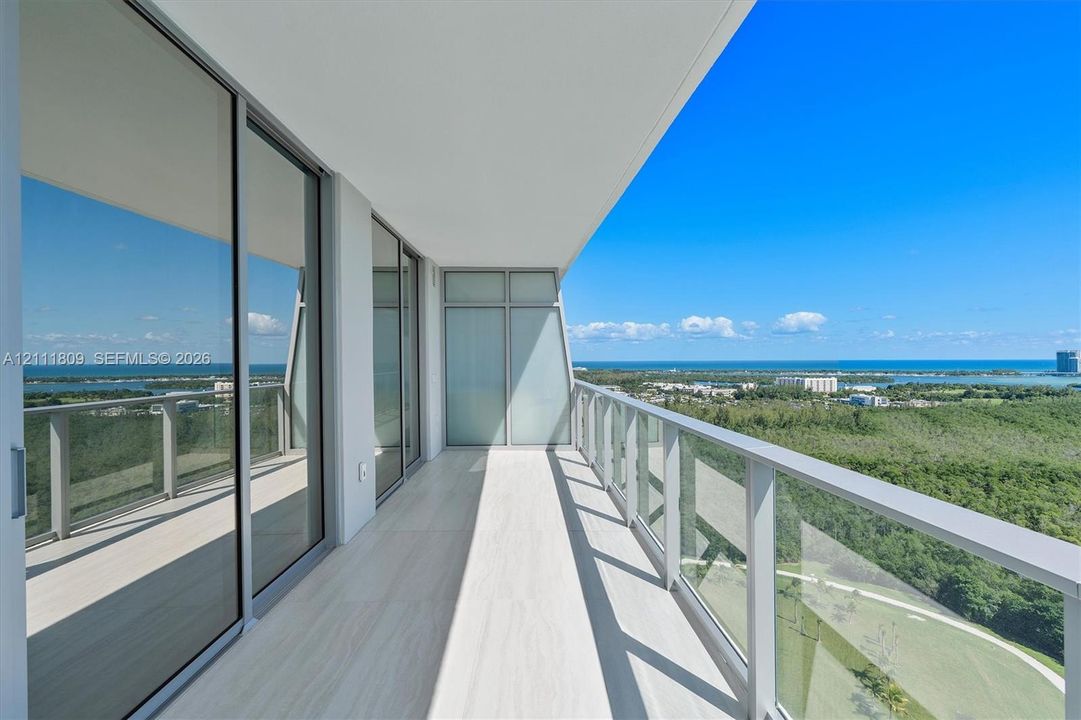 Covered balcony featuring tile flooring, full-height glass sliding doors, and a transparent glass railing overlooking a landscape with water in the distance