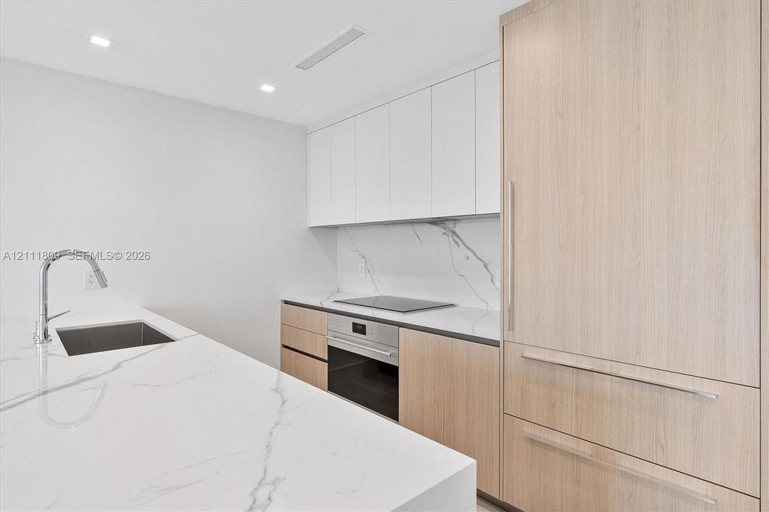 Kitchen featuring stone-finish countertops and backsplash, integrated paneled refrigerator, flat-panel wood-finish cabinetry, white upper cabinets, and a stainless steel undermount sink