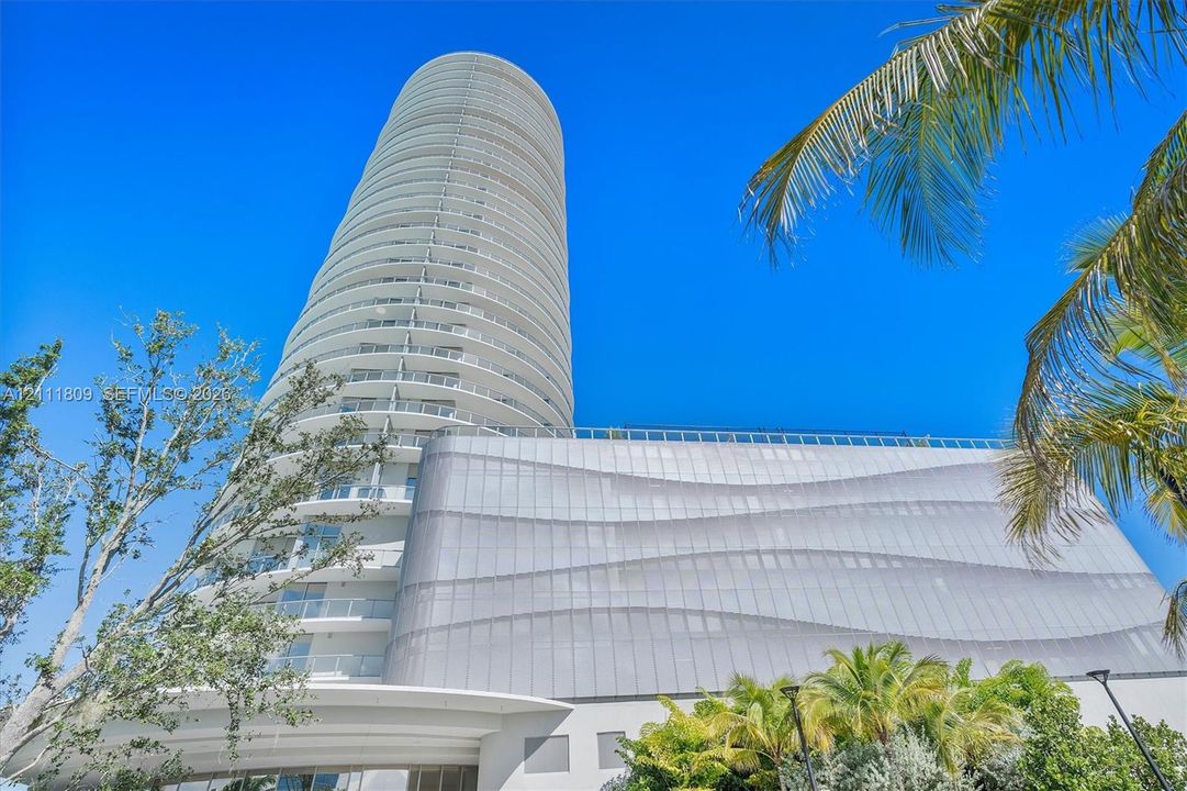 High-rise residential tower featuring a cylindrical glass architecture, wrap-around balconies, and a textured metal facade