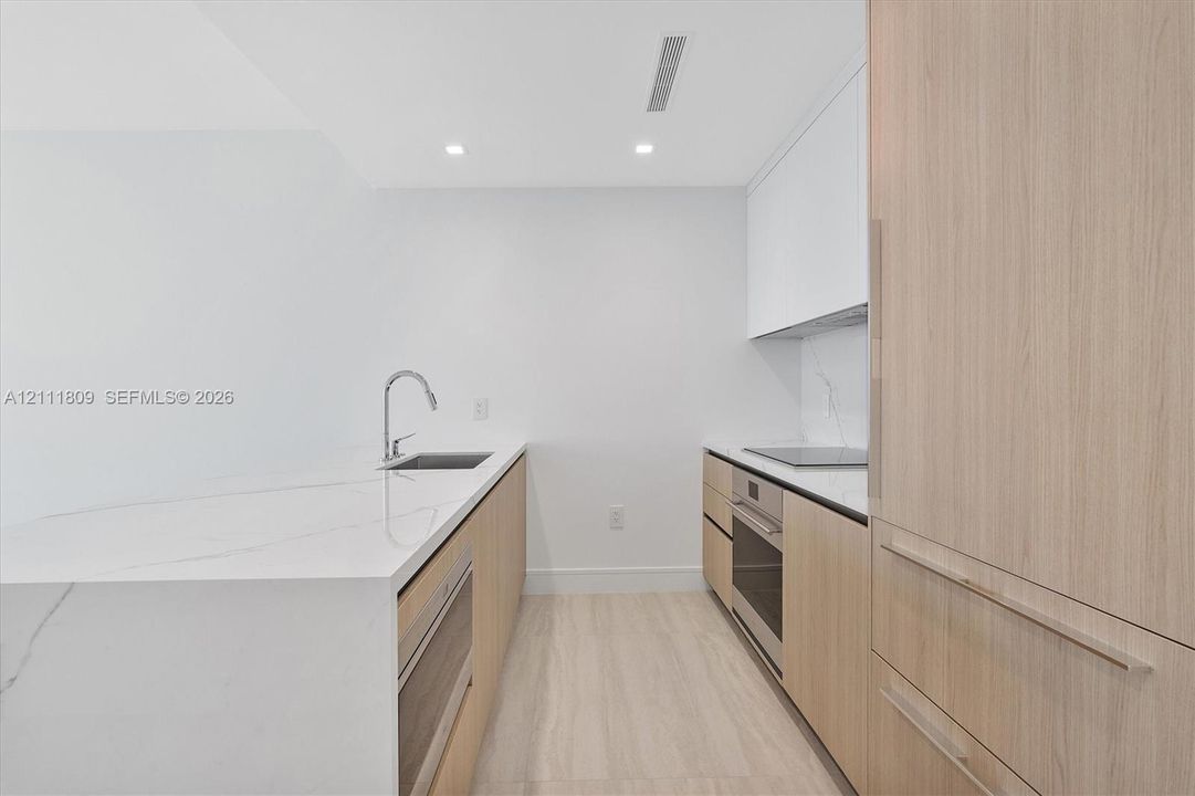 Kitchen featuring flat-panel wood cabinetry, white stone countertops with integrated backsplash, built-in stainless steel appliances, a gooseneck faucet, and light-toned flooring
