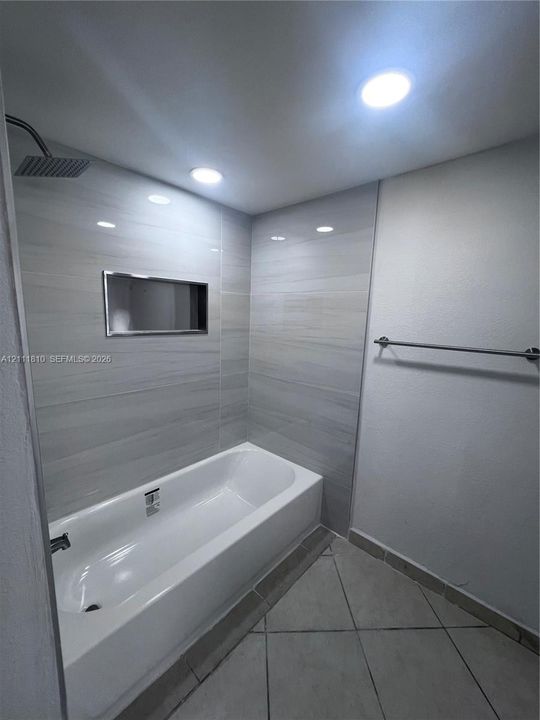 Bathtub with integrated shower, recessed tile niche, oversized rectangular wall tile, brushed metal hardware, and recessed ceiling lighting