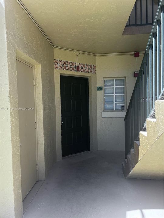 Covered entryway featuring a solid-panel entry door, decorative tile accent wall, single-pane window with security bars, and metal staircase
